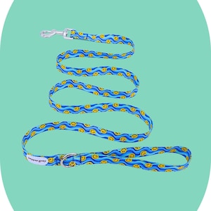 May include: A blue and yellow patterned dog leash with a repeating smiley face design. The leash has a silver metal clasp and a matching ring at the handle. The brand name "peppergrey" is printed on the leash.