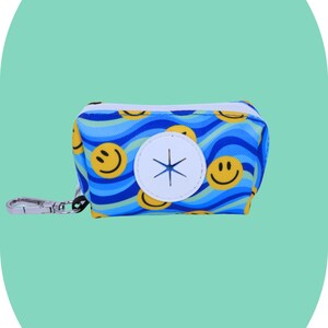 Puede incluir: Dispensador de bolsas para excrementos de perro con estampado azul y amarillo de caras sonrientes. Cuenta con una abertura circular blanca y un mosquetón plateado. Mide aproximadamente 10 cm de largo.