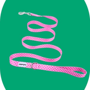May include: Pink dog leash with a checkered pattern. The leash has a silver-colored metal clasp and a matching ring. The brand name "pepper grey" is printed on the leash.
