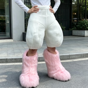May include: A pair of light pink, fluffy, faux fur boots. The boots are worn with white, textured shorts. The shorts have a high waist and a unique, rounded design. The outfit is worn outdoors on a paved surface.