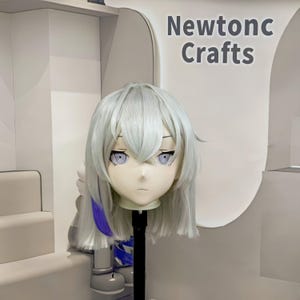Op de afbeelding: Een wit anime-stijl masker met lichtpaarse ogen en wit haar met een blauwe lok. Het masker is gemonteerd op een zwarte standaard. De tekst "Newtonc Crafts" is zichtbaar op de achtergrond.