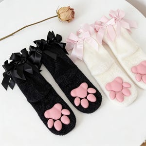 Peut inclure: Deux paires de chaussettes hautes, une noire et une blanche, avec des empreintes de pattes roses sur les semelles. Chaque chaussette est ornée d'un nœud en satin en haut. Présentées sur une surface blanche.