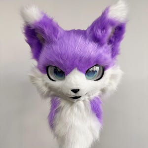 May include: A plush fox head with purple fur and white accents. The fox has large, expressive blue eyes and a black nose. The ears are pointed with white tips. The overall design is cute and cartoonish, suitable for cosplay or costume use.