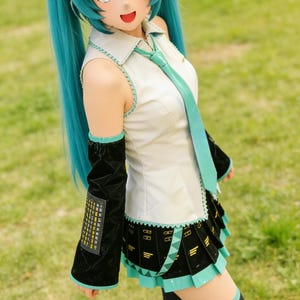 May include: A cosplayer in a teal wig and costume, resembling a character from a Japanese anime. The outfit includes a white top with a teal tie, a black skirt with teal accents, and black arm sleeves. The cosplayer is standing on green grass.
