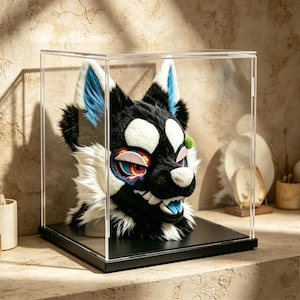 May include: A black and white furry wolf head with blue and white details, presented in a clear acrylic display case. The wolf head features white fur around the snout and a colourful eye design. The case rests on a black base.
