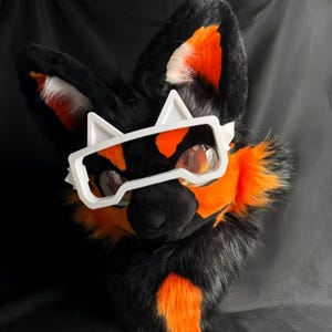 May include: A black and orange furry fox head with white glasses. The fox has large ears with orange accents and a black nose. The glasses are white and have a futuristic design. The fur is a mix of black, orange, and white.