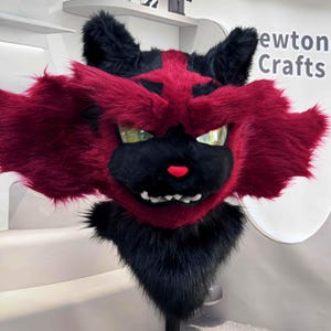 May include: A plush, red and black cat-like mask with a fierce expression. The mask features a black muzzle, ears, and neck fur, with red fur framing the face. The eyes are a reflective green, and the nose is red. The mask is labeled "Newton Crafts".