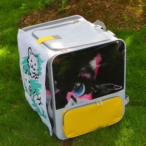 May include: A gray pet carrier with a yellow front pocket and a clear front panel. The carrier has a white side panel with cartoon dog illustrations. A black and white cat with pink accents is visible inside.