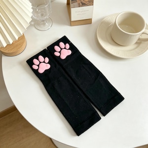 Op de afbeelding: Een paar zwarte armwarmers met een roze pootafdrukontwerp aan de bovenkant. De armwarmers zijn gemaakt van een zacht materiaal en worden op een wit oppervlak gepresenteerd.