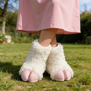 May include: White, fluffy sheep-style slippers with pink hoof-shaped toes. The slippers have a shaggy texture and are worn with a pink dress. The image is taken outdoors on a grassy lawn.