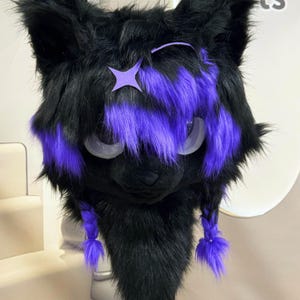 May include: A black cat-shaped plush headpiece with purple accents. The headpiece features purple streaks in the fur, a star-shaped detail, and braided pigtails. The cat has large, round, clear eyes.