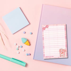 May include: A pastel-themed stationery set featuring a light blue notepad, a pink and white plaid notepad with a bow, and a teal pen. An ice cream cone eraser and scattered pastel-colored beads add a playful touch. The notepad reads "Believe in you."
