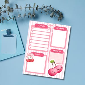 May include: A pink and white striped to-do list notepad with sections for reminders, notes, and priorities. The notepad features a cherry illustration and the words "TO DO LIST", "REMINDERS", "NOTES", and "PRIORITIES". A blue clipboard is in the background.