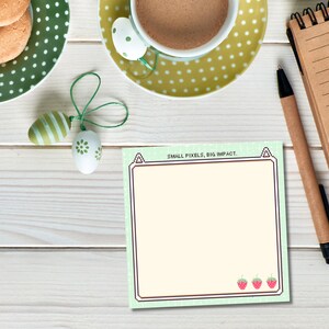 Puede incluir: Un bloc de notas verde y blanco con el texto "SMALL PIXELS, BIG IMPACT." El bloc de notas tiene un borde pixelado y tres fresas rojas en la parte inferior.