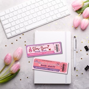 K&ouml;nnte beinhalten: Zwei rosafarbene und violette Ticket-f&ouml;rmige Lesezeichen mit dem Text "Slow Burn Book Club" und "Grumpy & Sunshine Book Club". Die Lesezeichen befinden sich auf einem wei&szlig;en Notizbuch mit einer wei&szlig;en Tastatur und rosa Blumen im Hintergrund.