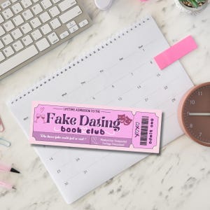 K&ouml;nnte beinhalten: Ein pinkfarbenes und violettes Ticket mit dem Text "Fake Dating Book Club" und dem Slogan "Who knew fake could feel so real?" Das Ticket hat ein pinkfarbenes Herz und eine Zeichnung von zwei Weingl&auml;sern.