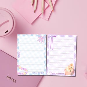 May include: Two lined notepads with a pastel color scheme. One notepad has a blue and white diamond pattern with a pink bow graphic and the text "Be bold & bright!". The other notepad has a purple and white diamond pattern with a ribbon graphic and a floral illustration.