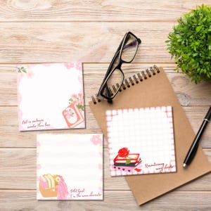 K&ouml;nnte beinhalten: Eine Sammlung quadratischer Haftnotizen mit floralen Designs und Texten, angeordnet auf einer Holzoberfl&auml;che. Ein Notizbuch, eine Brille, ein Stift und eine kleine Pflanze sind ebenfalls vorhanden. Die Notizen enthalten S&auml;tze wie "Lost in sentences sweeter than love."