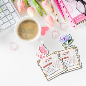 Op de afbeelding: Een bovenaanzicht van een witte bureau met een kop koffie, een toetsenbord, roze pennen en een notitieblok. Twee kleine kalenders voor juli en augustus 2026 zijn versierd met bloemen- en vlindermotieven. Roze tulpen en hartvormige paperclips maken het geheel af.