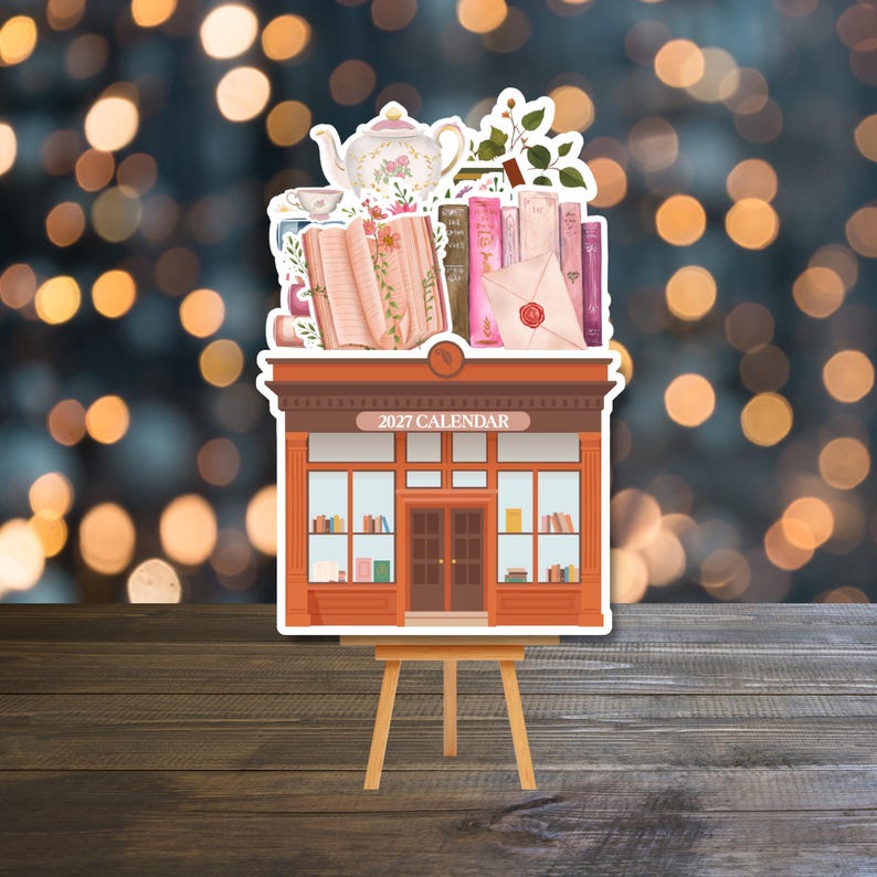 May include: A 2027 calendar featuring a storefront illustration topped with a collection of books, a floral teapot, a teacup, and a sealed envelope. The shop has a brown frame, glass windows, and a central door, all displayed on a wooden easel.
