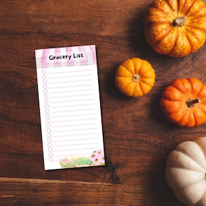 May include: A pink and white striped grocery list notepad with a floral design at the bottom. The notepad has a title that says "Grocery List". There are 25 blank lines with circles for checking off items. The notepad is on a brown wooden surface with four pumpkins.