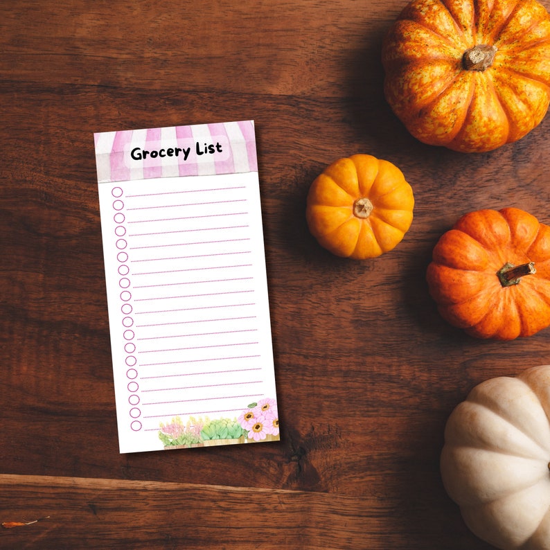 May include: A pink and white striped grocery list notepad with a floral design at the bottom. The notepad has a title that says "Grocery List". There are 25 blank lines with circles for checking off items. The notepad is on a brown wooden surface with four pumpkins.