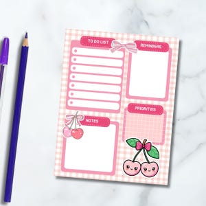 May include: Pink and white gingham patterned notepad with sections for a to-do list, reminders, notes, and priorities. Adorned with cherry illustrations and bows. A purple pen and blue pencil are visible.