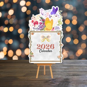 Op de afbeelding: Een decoratieve kalender voor 2026 op een klein houten ezel. De kalender heeft een vlinderontwerp en bloemaccenten. De bovenkant van de kalender is versierd met kleurrijke vlinders en bloemen. De tekst "2026 Calendar" wordt op de kalender weergegeven.