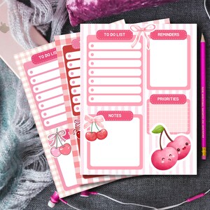 May include: Pink and white to-do list notepads with cherry illustrations. The notepads feature sections for to-do lists, reminders, notes, and priorities. A pink pencil and earbuds are also visible.