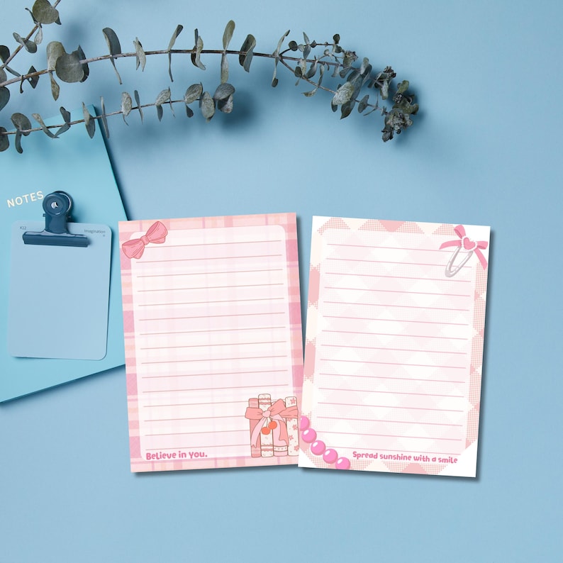 May include: Two pink notepads with lined pages, decorated with bows and gift illustrations. One notepad has the text "Believe in you." and the other says "Spread sunshine with a smile." A blue notepad with a clip is in the background.