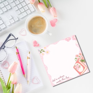 K&ouml;nnte beinhalten: Eine Draufsicht mit einer wei&szlig;en Tastatur, einer Kaffeetasse und einem Notizblock mit rosa floralen Akzenten. Zwei Stifte, eine Brille und herzf&ouml;rmige Dekorationen sind ebenfalls vorhanden. Der Notizblock tr&auml;gt den Text "Lost in sentences sweeter than love."