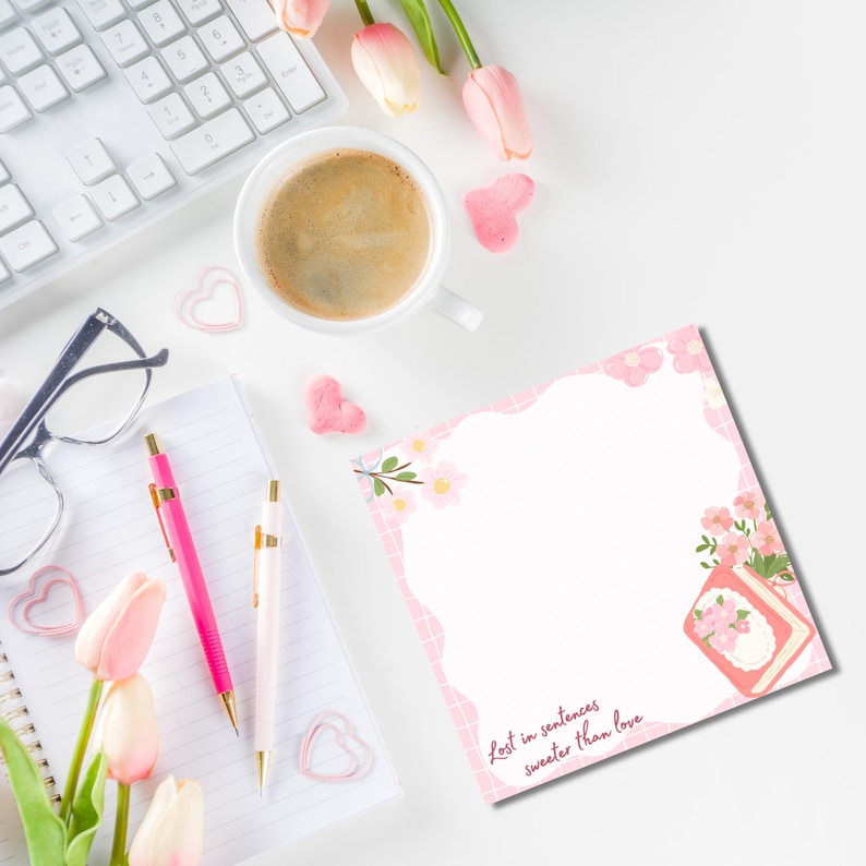 K&ouml;nnte beinhalten: Eine Draufsicht mit einer wei&szlig;en Tastatur, einer Kaffeetasse und einem Notizblock mit rosa floralen Akzenten. Zwei Stifte, eine Brille und herzf&ouml;rmige Dekorationen sind ebenfalls vorhanden. Der Notizblock tr&auml;gt den Text "Lost in sentences sweeter than love."
