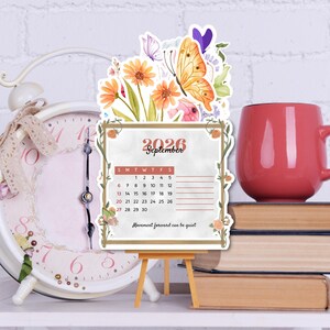 Op de afbeelding: Decoratieve september 2026 kalender met bloemen- en vlinderaccenten, gepresenteerd op een klein houten ezel. De kalender heeft een maandelijkse indeling met de tekst "Movement forward can be quiet."