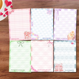 May include: Six lined notepads with pastel backgrounds and decorative borders. Each notepad features a bow, text, and other embellishments. The text includes phrases like "Believe in you" and "You are delightful."