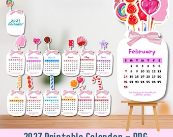 May include: A 2027 printable calendar set featuring twelve monthly mason jar-shaped cards decorated with pink bows and various candy illustrations. Each card displays the month name, calendar grid, and small text. A large February card sits on a wooden easel.