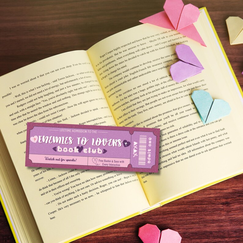 K&ouml;nnte beinhalten: Ein pinkfarbenes und violettes Ticket mit dem Text "Enemies to Lovers Book Club" und "Watch out for sparks!" Das Ticket liegt auf einem ge&ouml;ffneten Buch mit rosa, violetten und blauen Origami-Herzen.