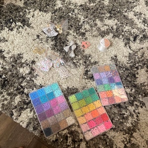 May include: Three clear plastic containers filled with colorful round beads. The containers are on a gray and white patterned rug. There are also two small bags of beads, a pair of scissors, and a small container of clear glue.