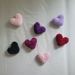 May include: Six heart-shaped pillows in various colors, including pink, purple, red, and black. The pillows are made of a soft, fuzzy material and are arranged on a white surface.