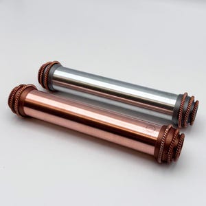 Op de afbeelding: Twee cilindrische metalen buizen, één met een gepolijste koperen afwerking en de andere met een geborstelde zilveren afwerking. Beide buizen hebben donkere, getextureerde eindkappen met een touwachtig patroon en een klein zonmotief.