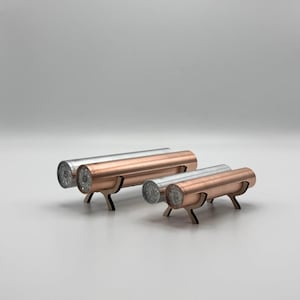 Peut inclure: Quatre bougeoirs cylindriques en métal. Deux sont argentés et deux sont de couleur cuivre. Chaque support a une base en bois à trois pieds. Les bougeoirs sont disposés en rangée sur une surface blanche.