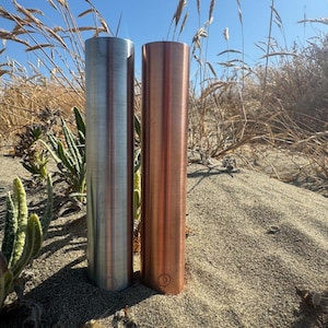 Puede incluir: Dos tubos cilíndricos de metal colocados verticalmente en la arena. Un tubo tiene un acabado plateado cepillado y el otro un acabado de cobre pulido. Cada tubo presenta un pequeño icono de sol grabado cerca de la base.