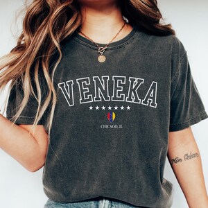 May include: A dark grey t-shirt with a white graphic that says "VENEKA" with stars and the text "CHICAGO, IL". The model is wearing blue jeans and white trainers.