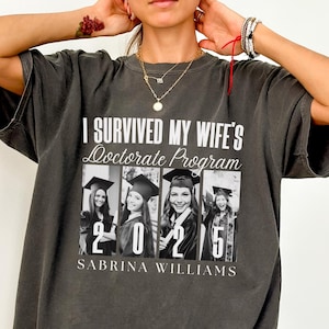 May include: A dark gray t-shirt with the text "I survived my wife's doctorate program 2025 Sabrina Williams" and a photo of four people in graduation caps.