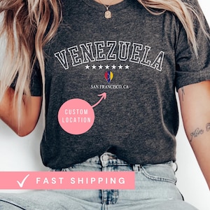 May include: Dark grey heather t-shirt with the word "VENEZUELA" in white arched lettering. Below the word "VENEZUELA" are five white stars, a graphic, and the text "SAN FRANCISCO, CA". The shirt also has the text "CUSTOM LOCATION".