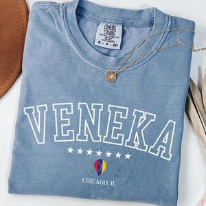May include: A light blue t-shirt with the word "Veneka" printed in white with a white outline. The shirt also has a small graphic of a heart with the flag of Romania inside and the text "Chicago, IL" printed below.