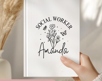 May include: White notebook with black text that reads "Social Worker" in a circle with a floral design and butterflies. The name "Amanda" is written below the circle. The notebook is a personalized gift.
