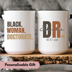 May include: Two white ceramic mugs with black handles. The mug on the left has the text "BLACK. WOMAN. DOCTORATE." in brown, black, and beige lettering. The mug on the right has the text "DR. Johnnie Ph.D. 2024" in brown, black, and beige lettering.