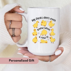 May include: White ceramic mug with a handle, featuring a design of yellow cartoon ducks and the text "My Ducks don't even know what a row is." The mug is held by a person wearing a white sweater.