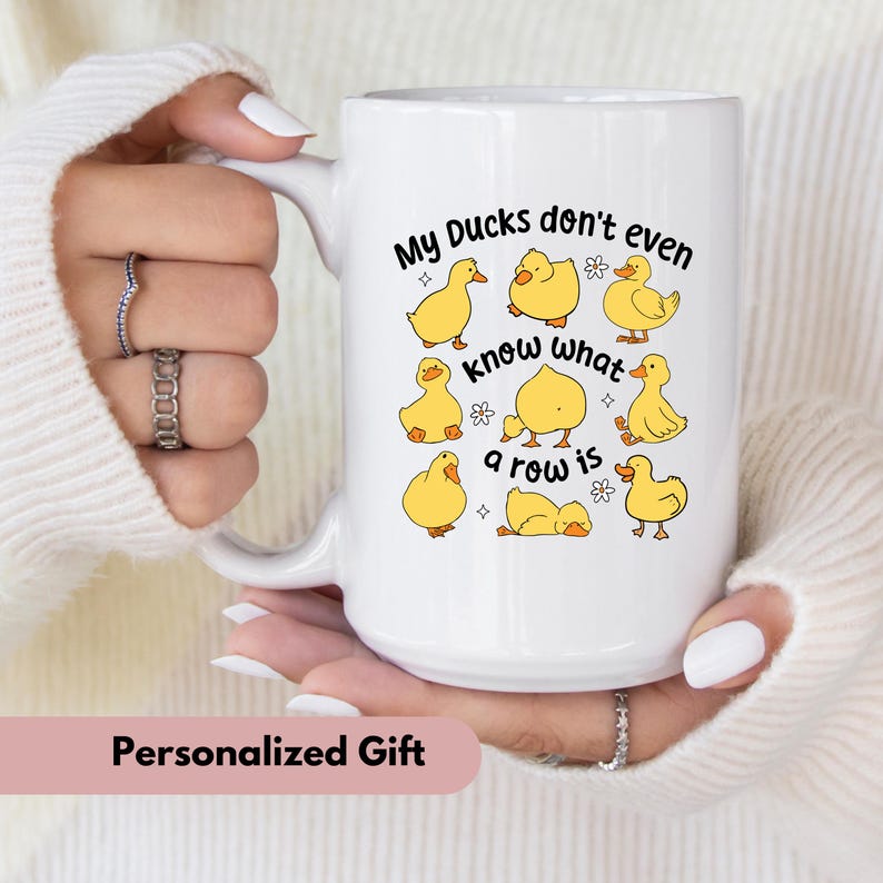 May include: White ceramic mug with a handle, featuring a design of yellow cartoon ducks and the text "My Ducks don't even know what a row is." The mug is held by a person wearing a white sweater.