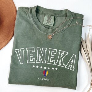 May include: A green t-shirt with the word "VENEKA" in white outlined letters, five white stars, and a small graphic of a flag. The text "CHICAGO, IL" is printed below the graphic. The t-shirt is folded and laid on a white surface.